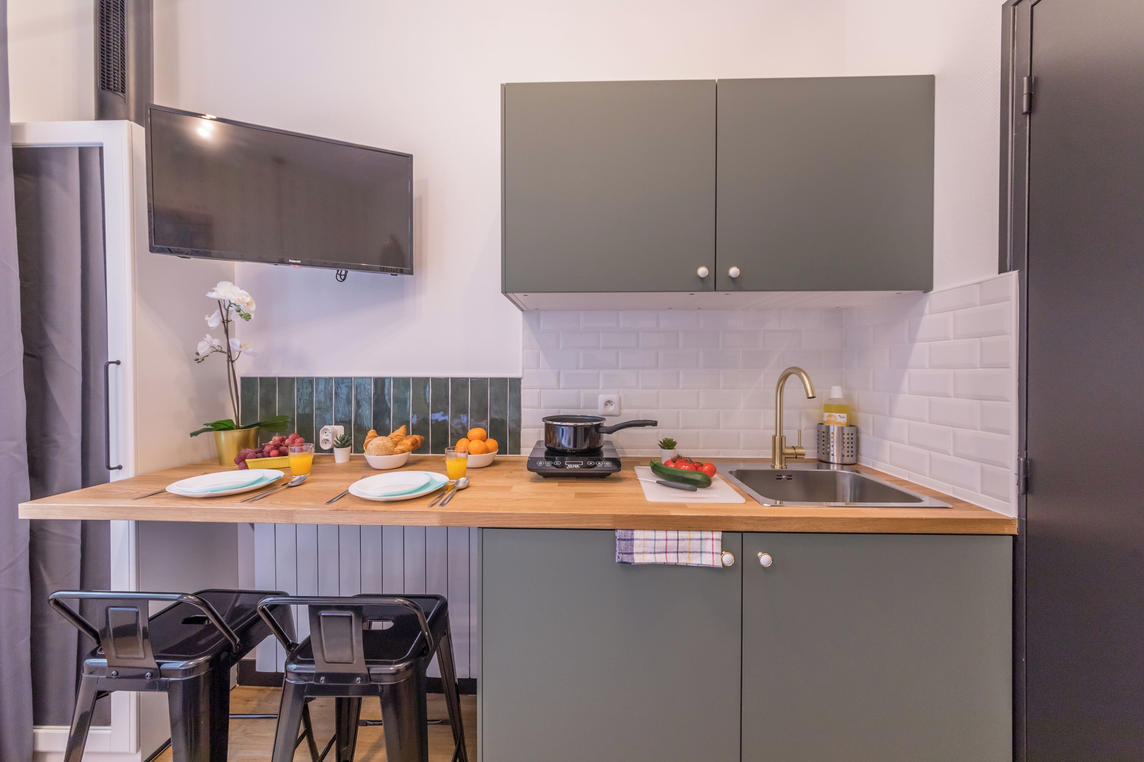 A modern kitchen with a wooden countertop, two black bar stools, a sink, and a television mounted above the counter.