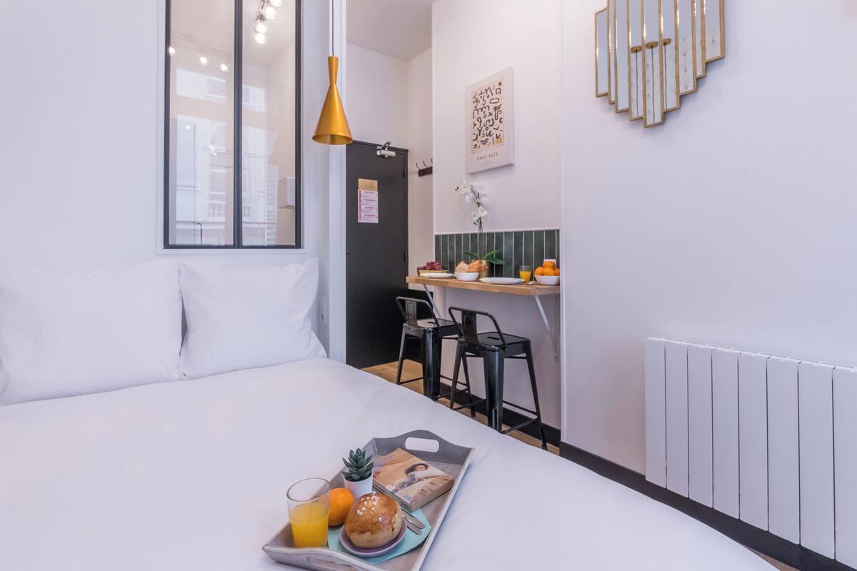 A hotel room with a bed, a breakfast tray, a small table with chairs, a refrigerator, and a wall-mounted light fixture.