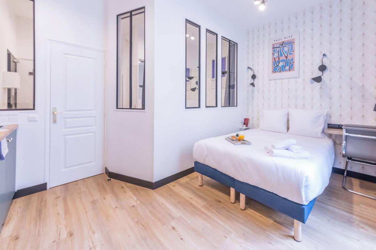 A modern hotel room with a wooden floor, a bed with white linens, a bedside table, a mirror, and a framed artwork on the wall.