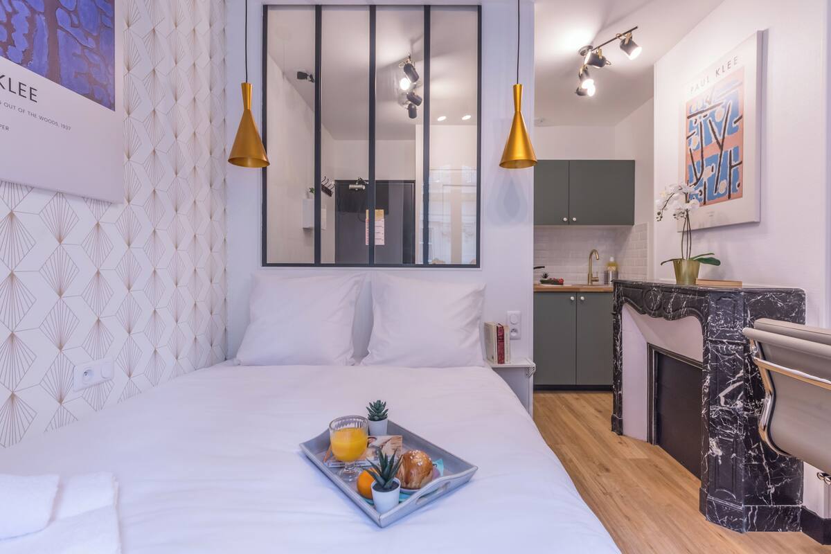 A hotel room with a bed, a breakfast tray, a small potted plant, and a view of the kitchen.