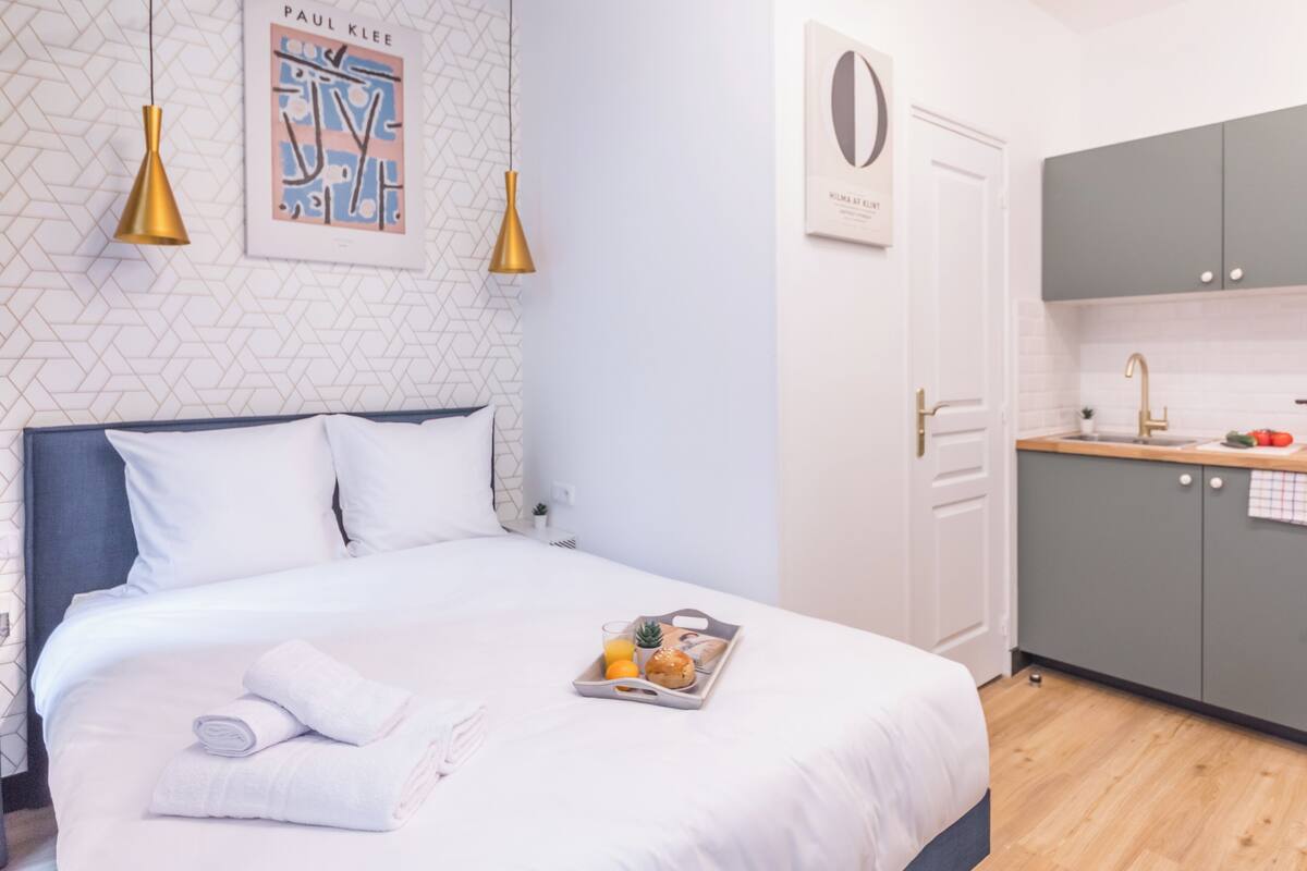A neatly made bed with a tray of breakfast items, a wall with geometric patterns, and a kitchen with grey cabinets.