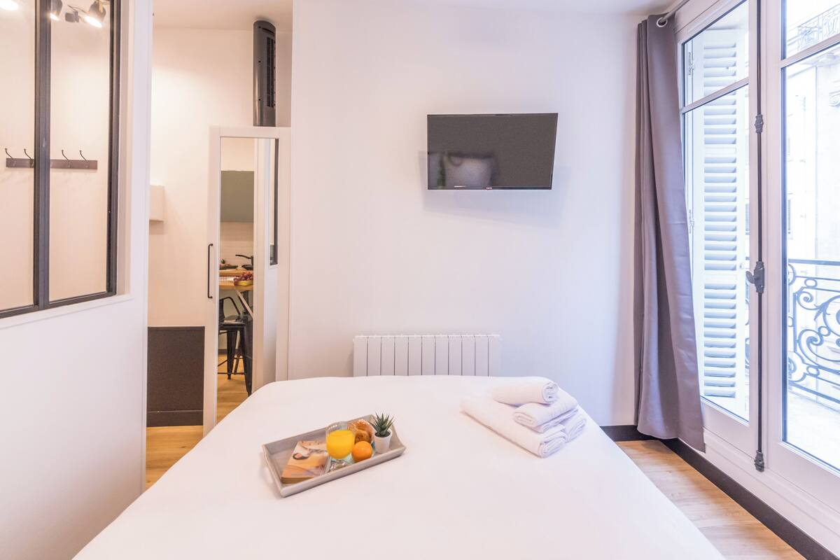 A hotel room with a bed, a tray with breakfast items, a TV on the wall, and a window with blinds.