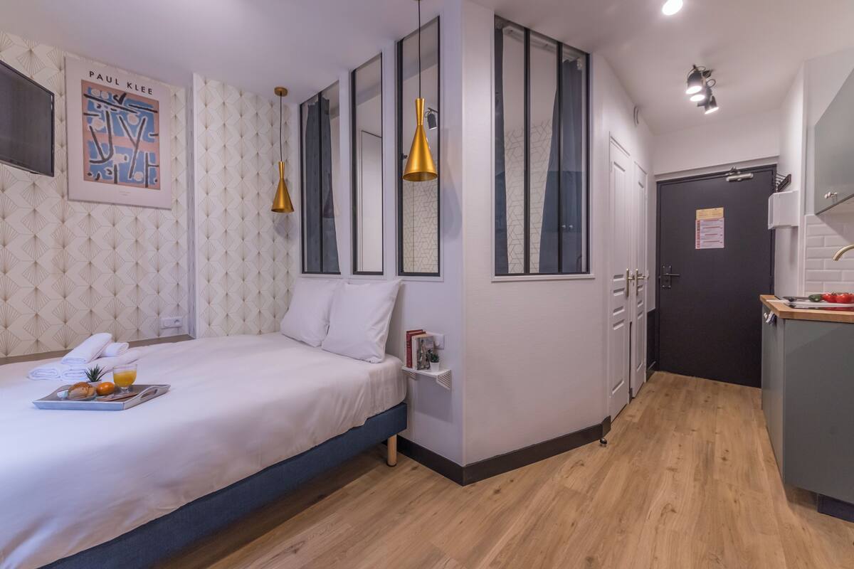 A modern hotel room with a neatly made bed, a small kitchenette, and a large window.