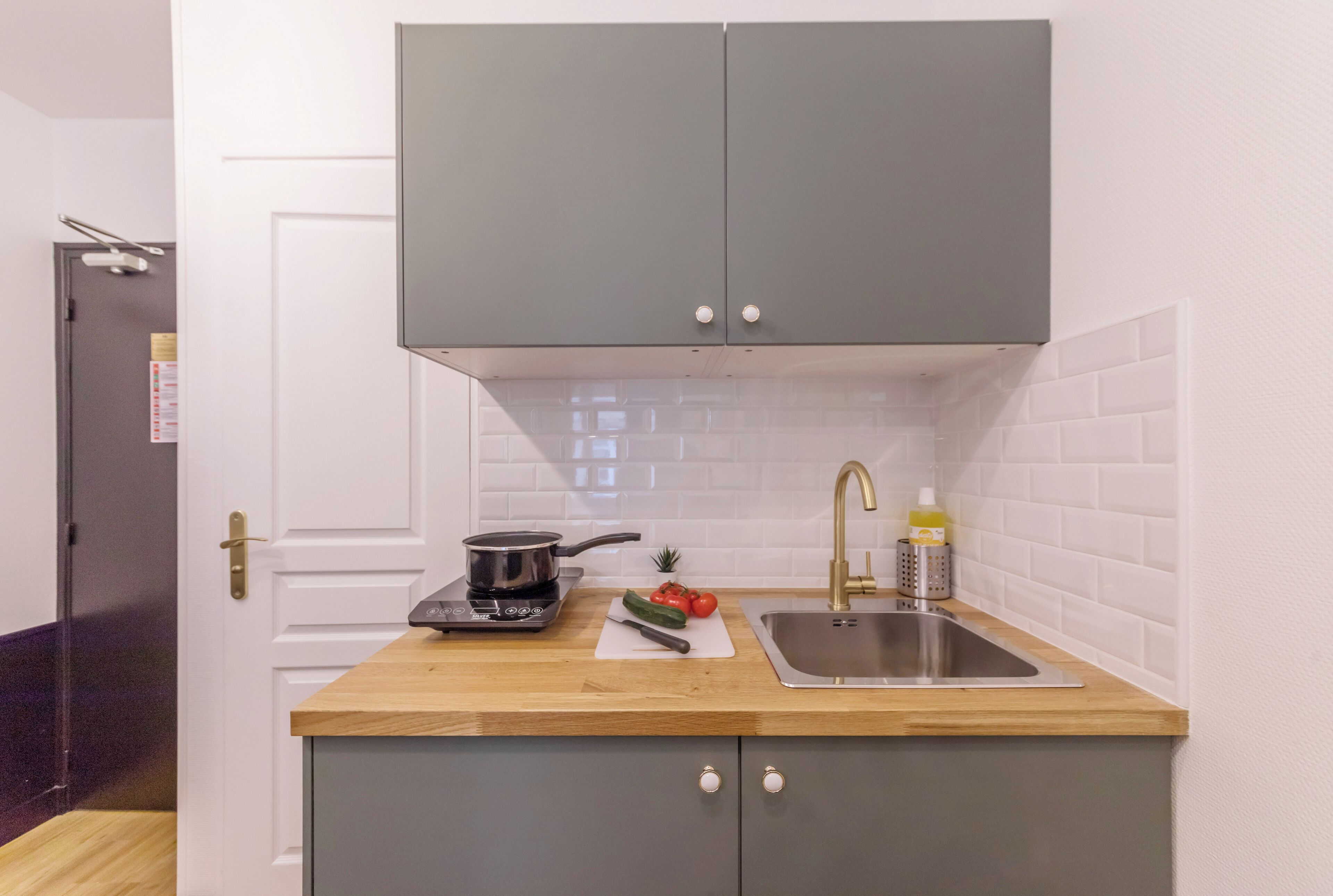 Une cuisine compacte avec des meubles gris, un plan de travail en bois, une cuisinière encastrée, un évier et une petite fenêtre.