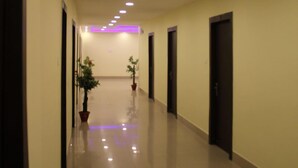 A long, well-lit hallway with doors on both sides, potted plants, and a tiled floor. - Hotel Vibrant Lawn And Banquet (Danapur)