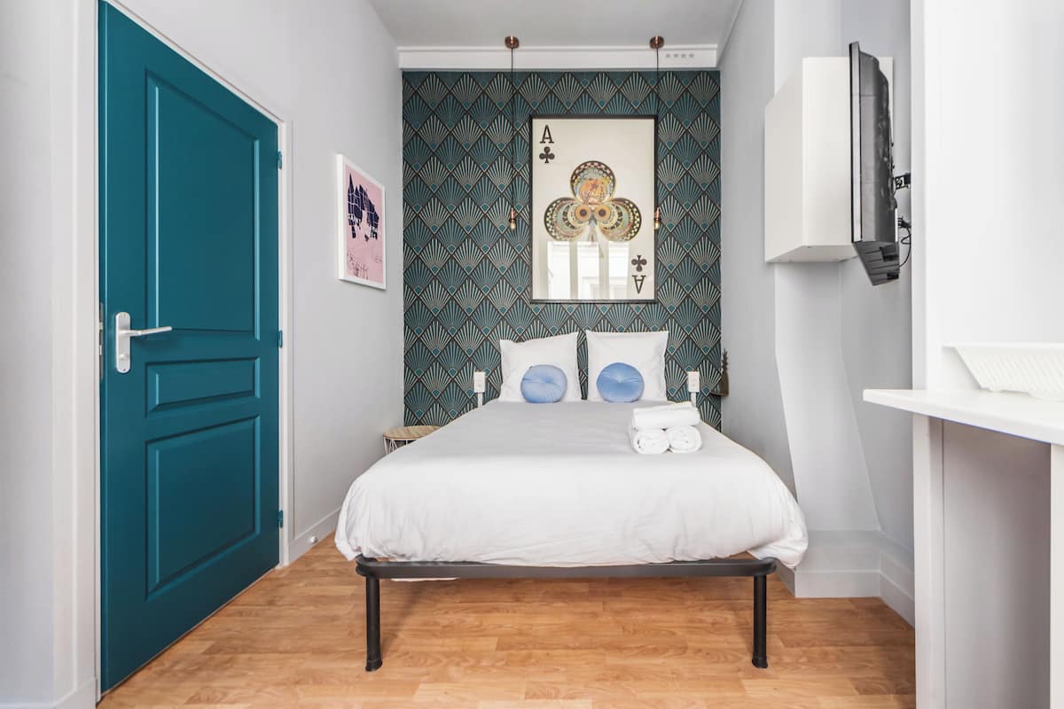 Une chambre avec une porte de couleur turquoise, un papier peint à motifs, un lit avec une literie blanche et un téléviseur fixé au mur.