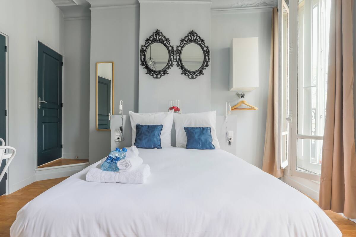 Une chambre d’hôtel avec un grand lit, deux miroirs, une table de chevet, une lampe et une fenêtre avec des rideaux.