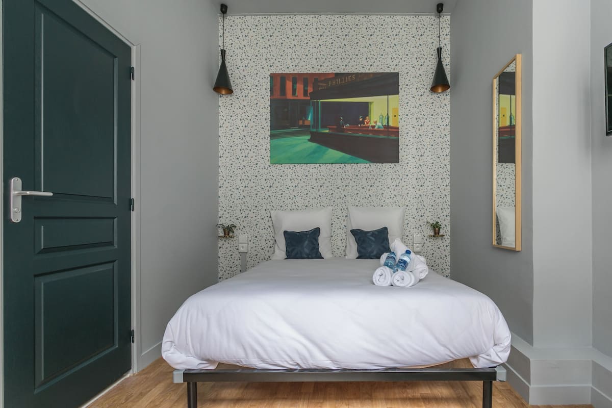 Un lit bien fait, avec du linge de lit blanc et deux oreillers, un tableau représentant un paysage urbain au-dessus, et un miroir accroché au mur.