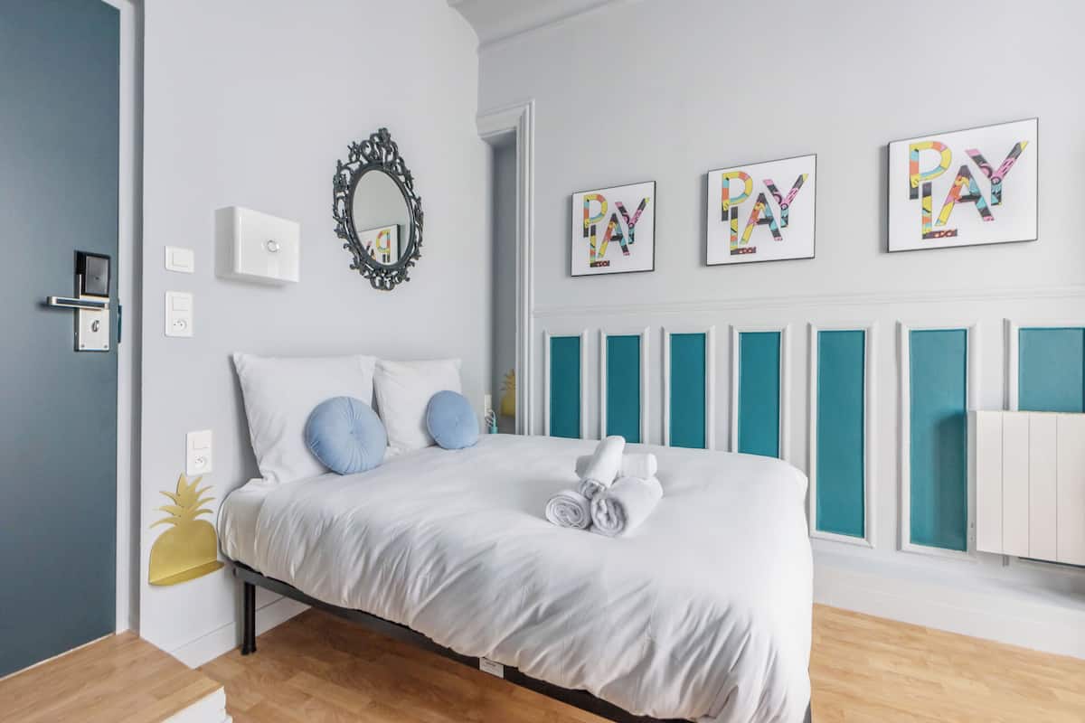 Une chambre moderne avec un lit, du linge de lit blanc, un miroir et trois œuvres d’art intitulées « PAY » accrochées au mur.
