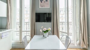 A dining area with a white table, two chairs, a vase with flowers, a flat-screen TV mounted on the wall, and a window with curtains. - WS Saint-Lazare - Opera (Paris)