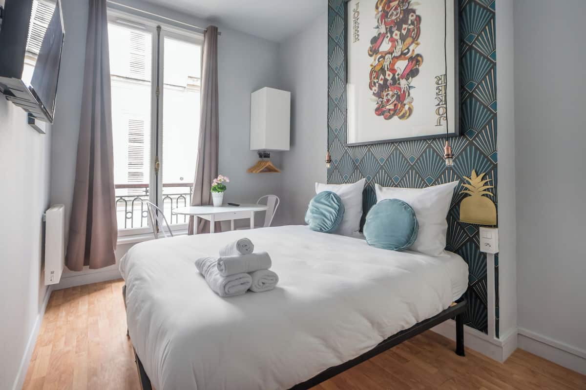 Une chambre d’hôtel comprenant un lit, un bureau, une chaise, une fenêtre avec des rideaux et un mur orné d’un motif géométrique.