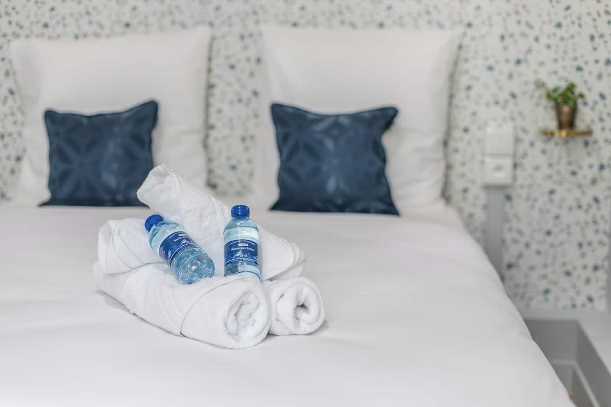 Un lit bien fait, avec des serviettes blanches pliées en rouleau et deux bouteilles d’eau.