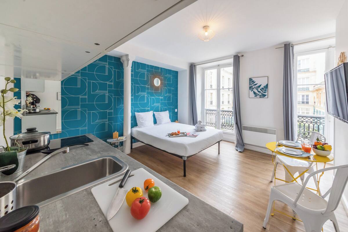 A modern hotel room with a bed, a kitchen area, and a balcony with a view of the city.
