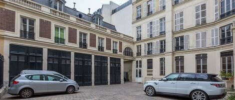 A courtyard with two parked cars, a building with multiple windows and balconies, and a brick pavement.