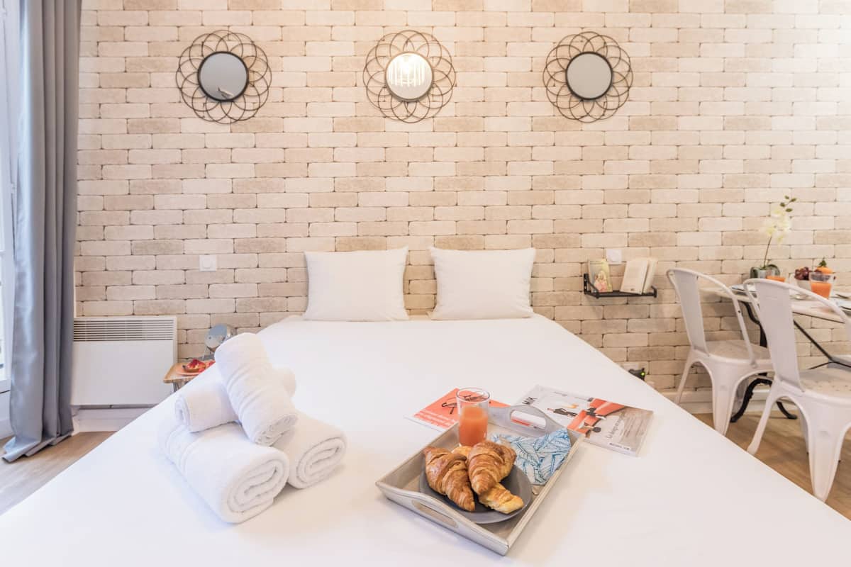 A hotel room with a brick wall, a bed with white linens, a tray with croissants and juice, and a desk with chairs.
