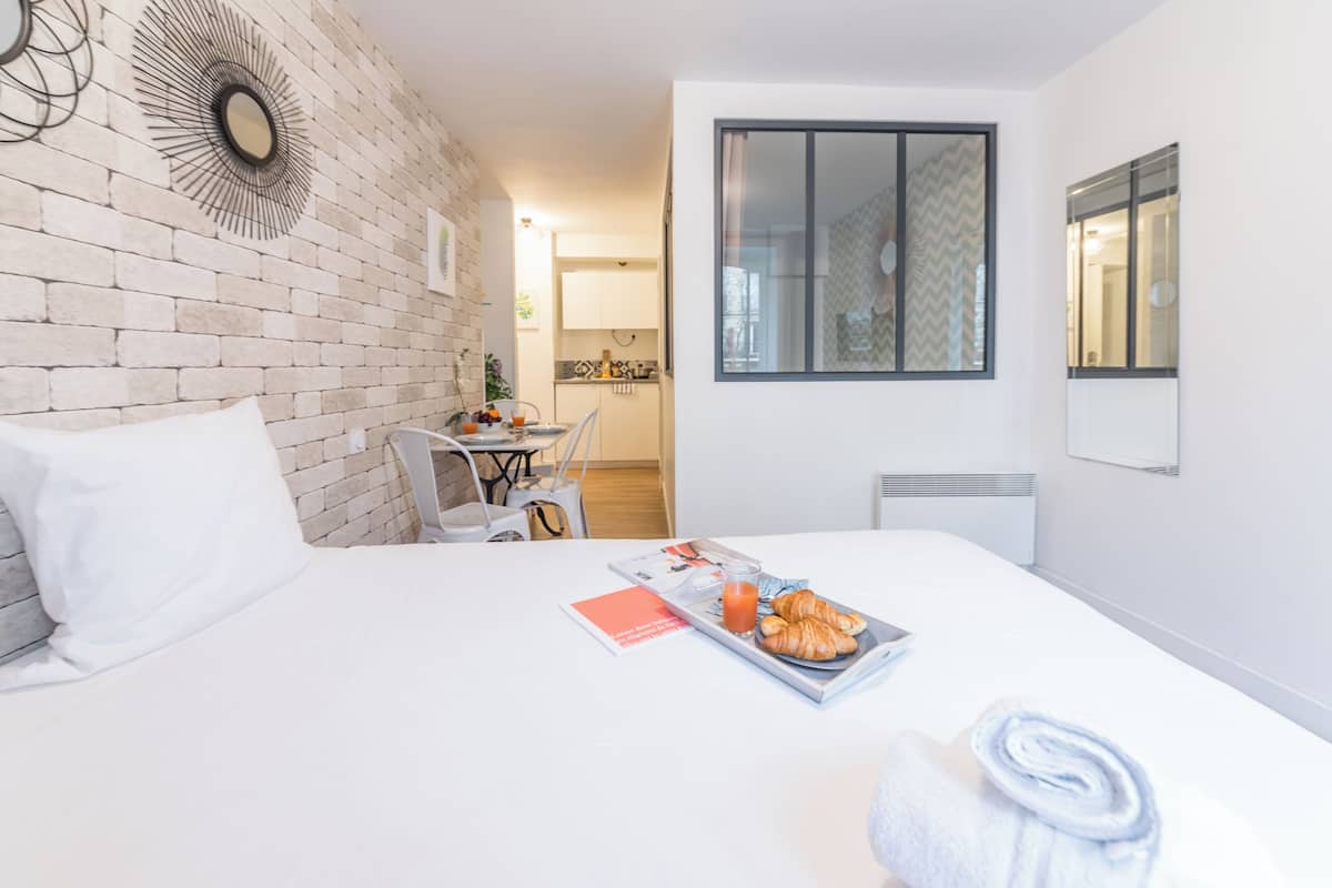 A hotel room with a brick wall, a bed with white bedding, a breakfast tray with croissants and juice, and a window with a view of the kitchen and hallway.