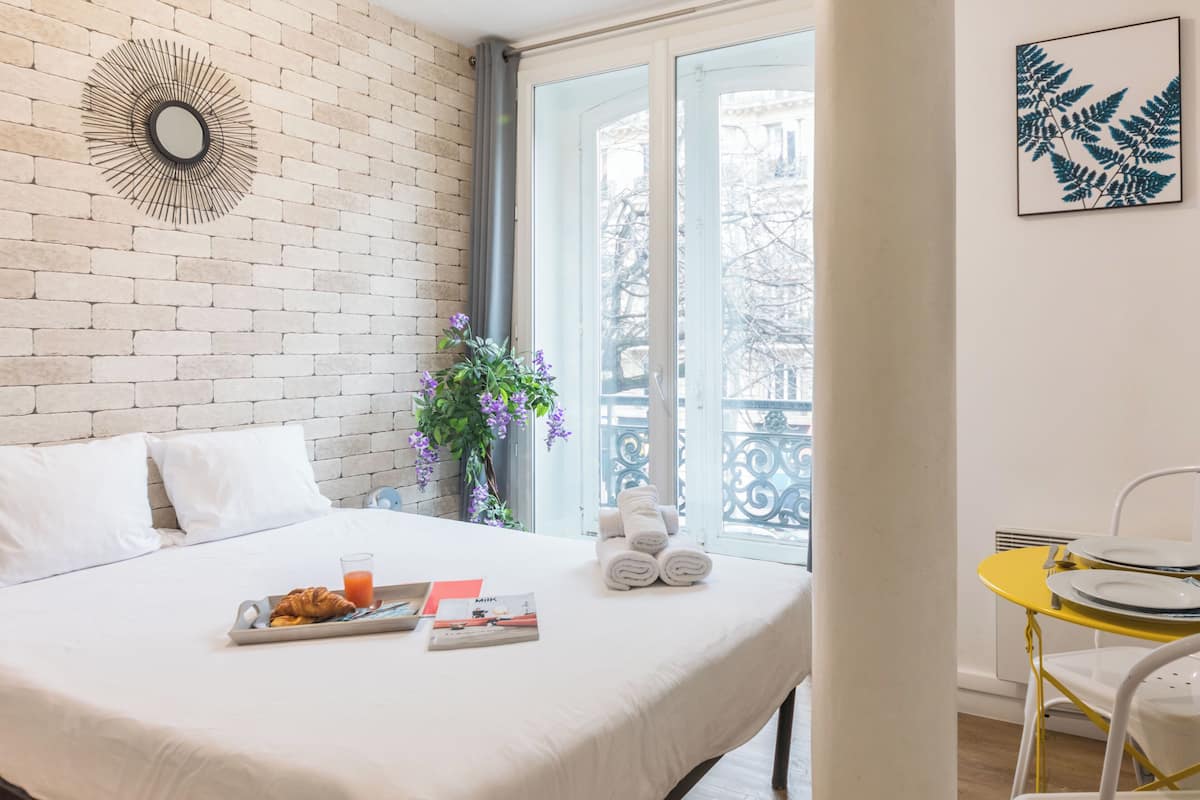 A hotel room with a bed, a breakfast tray, a window with a view, a plant, and a yellow table with plates.