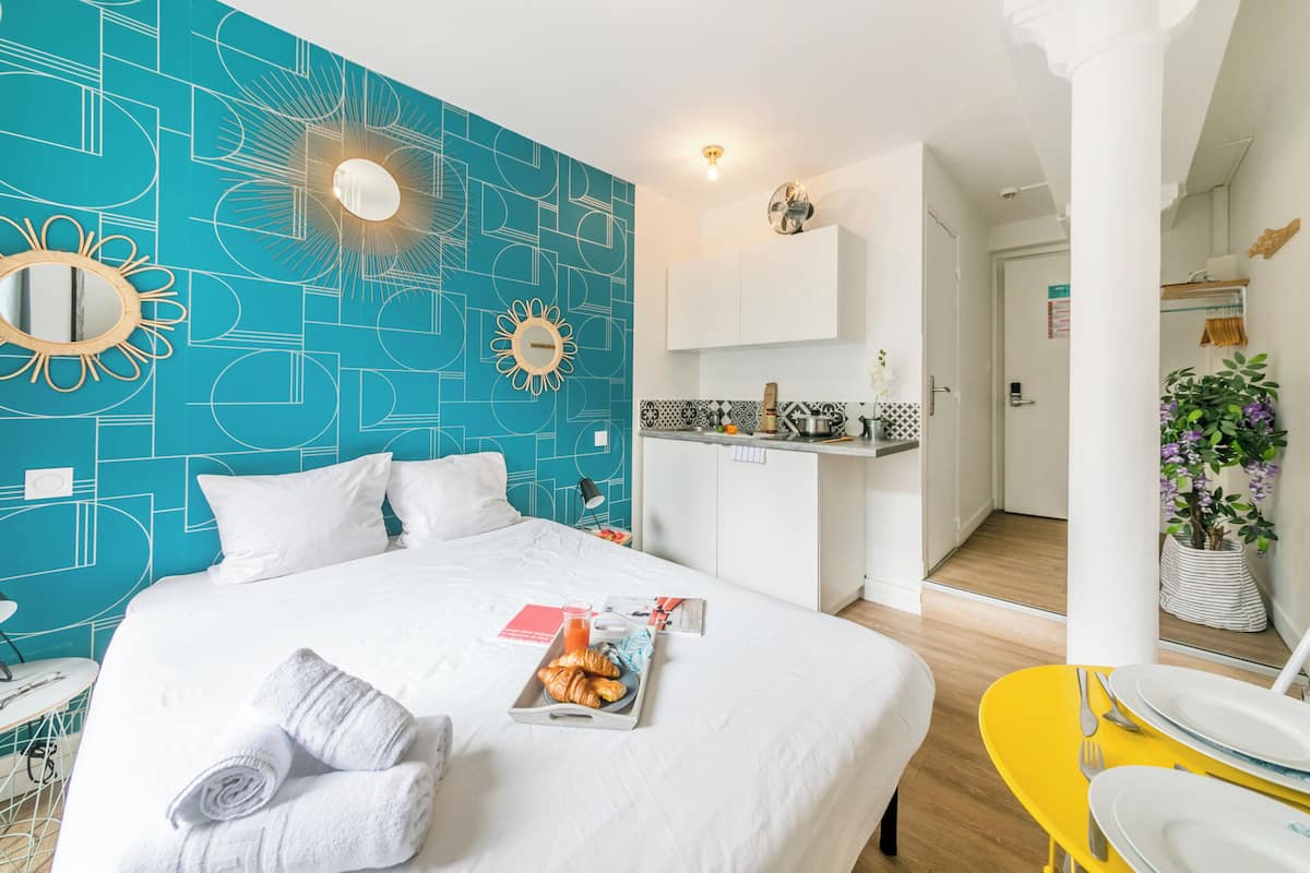 A modern hotel room with a geometric blue wall, a white bed, a breakfast tray, and a yellow side table.