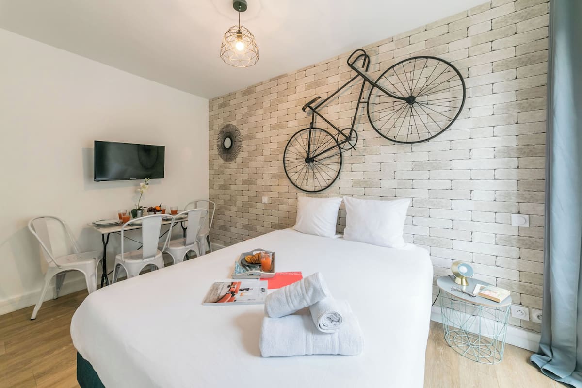 A hotel room with a large bed, a dining table with chairs, a television, and a bicycle mounted on the brick wall.