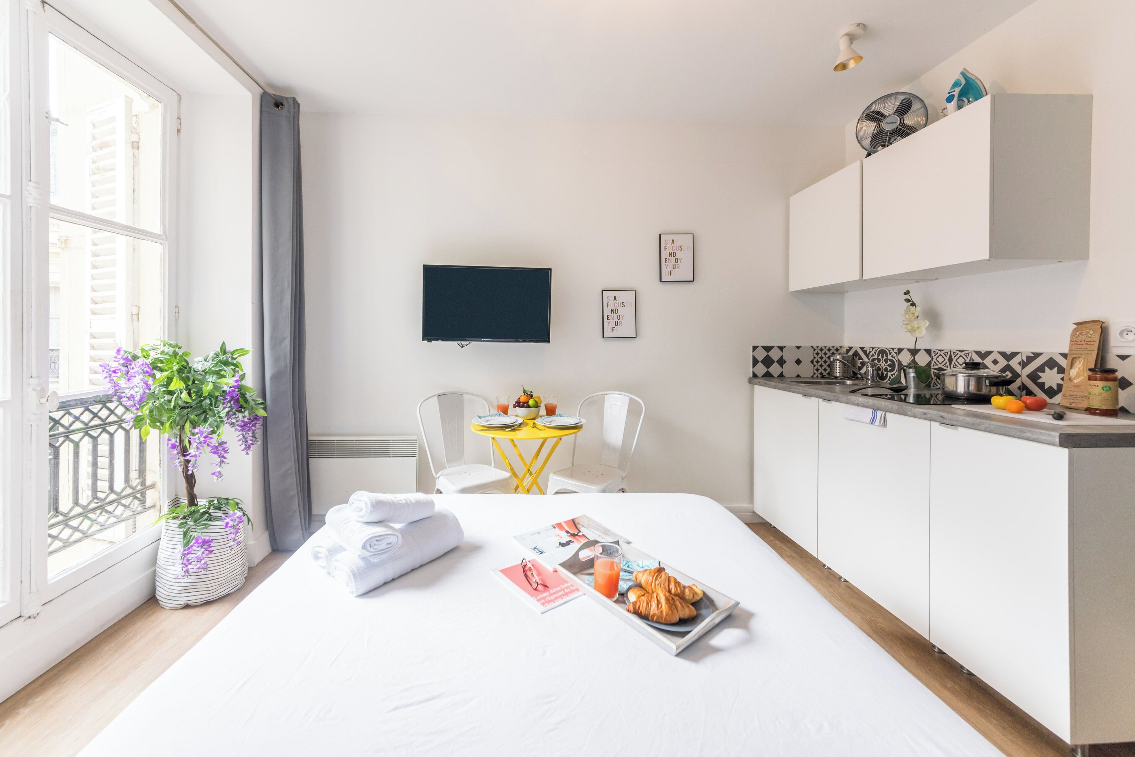A modern hotel room with a bed, a small dining area, a TV, and a kitchen area.