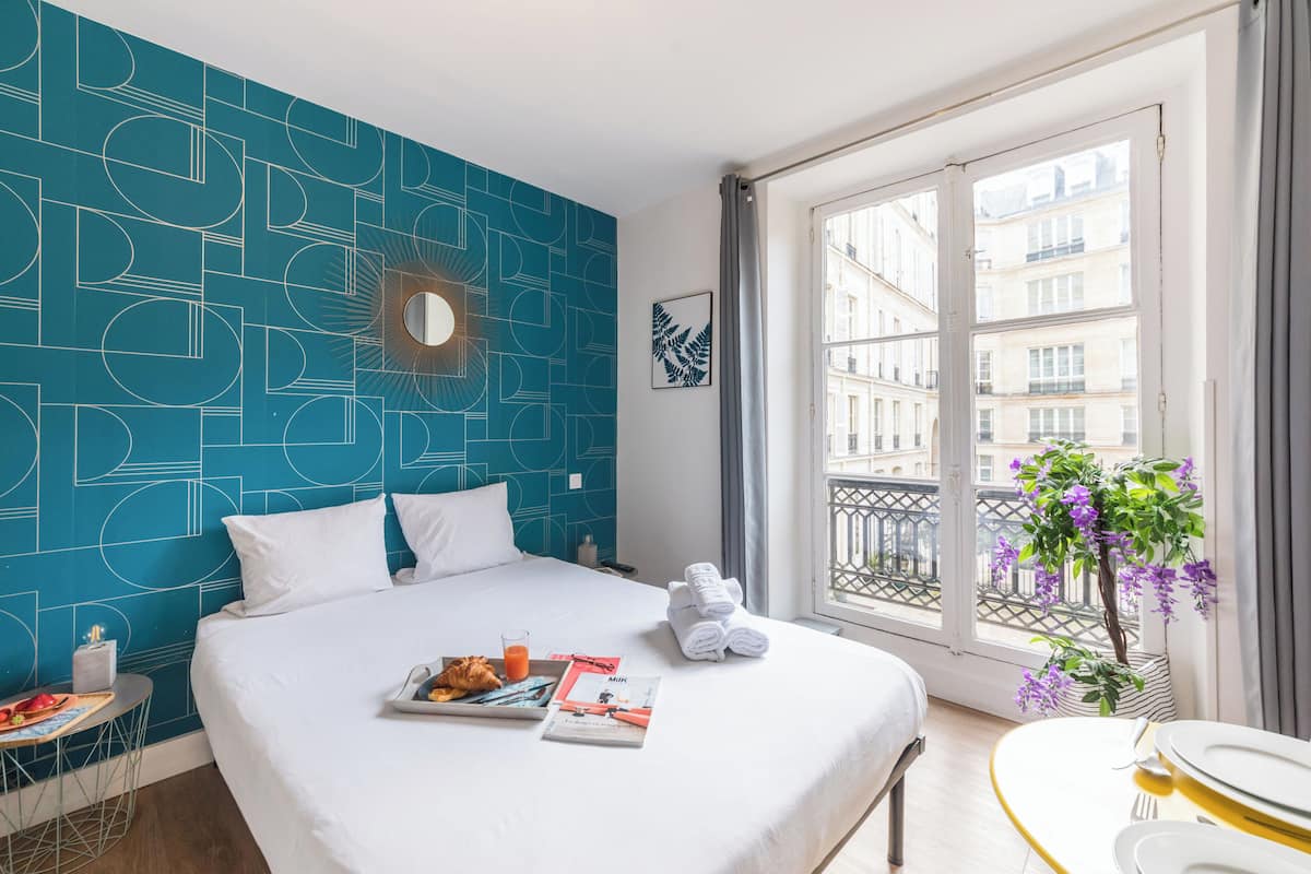 A hotel room with a large bed, a small table with breakfast, a window with a view of buildings, and a decorative wall.