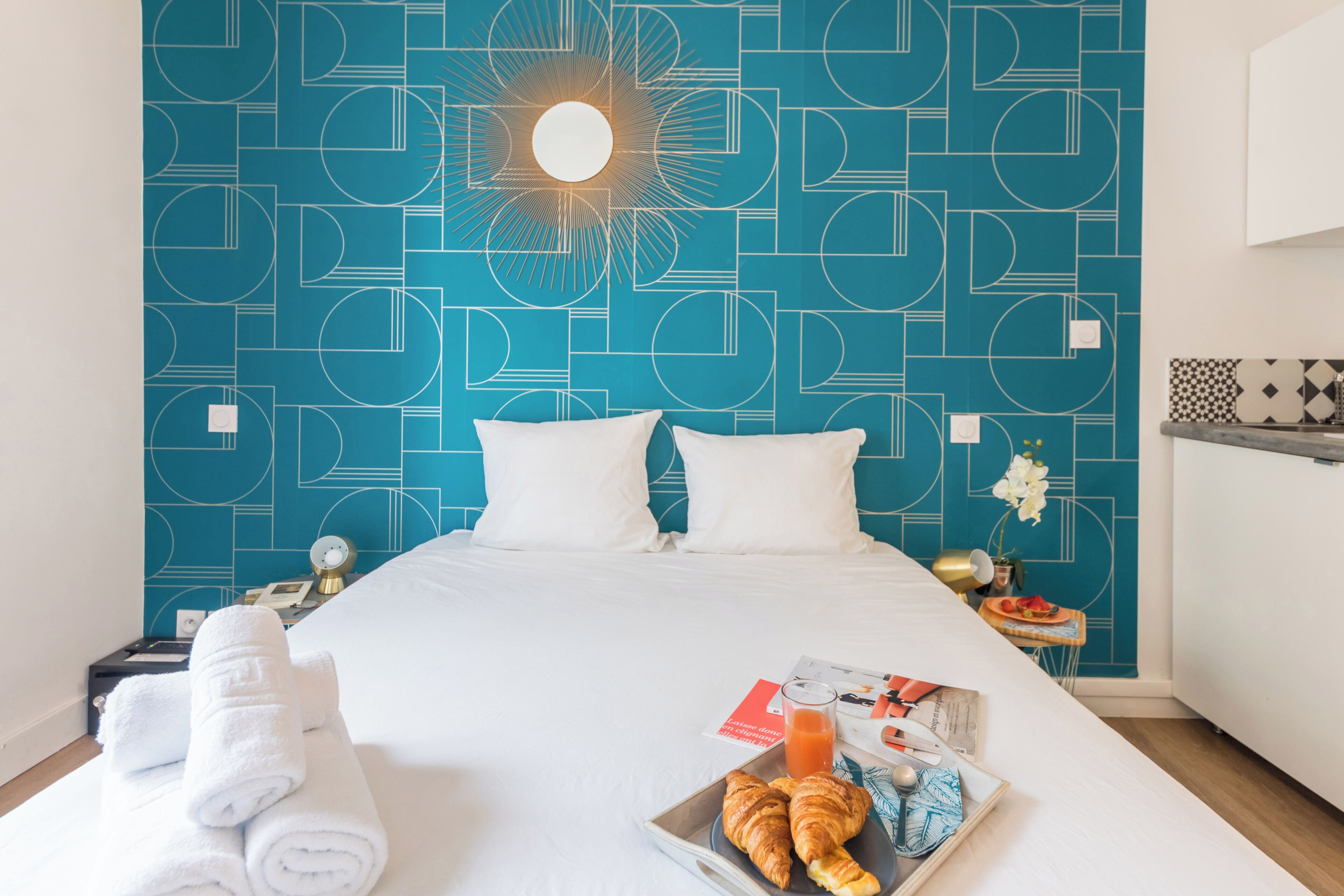 Une chambre d’hôtel avec un mur orné d’un motif géométrique, un lit avec du linge de lit blanc, deux oreillers, un plateau avec des croissants et un verre de jus d’orange, et une petite table avec une horloge et une plante.