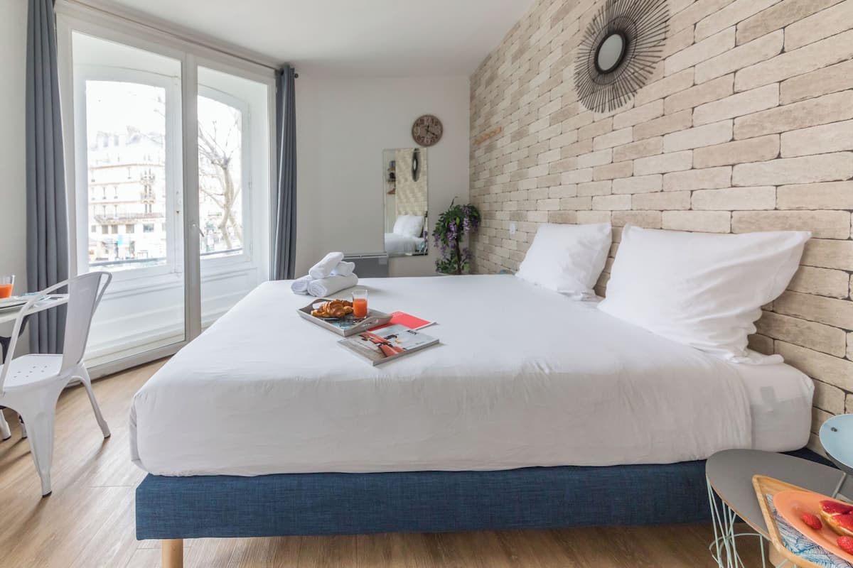 A modern hotel room with a large bed, a desk, and a view of the city through large windows.