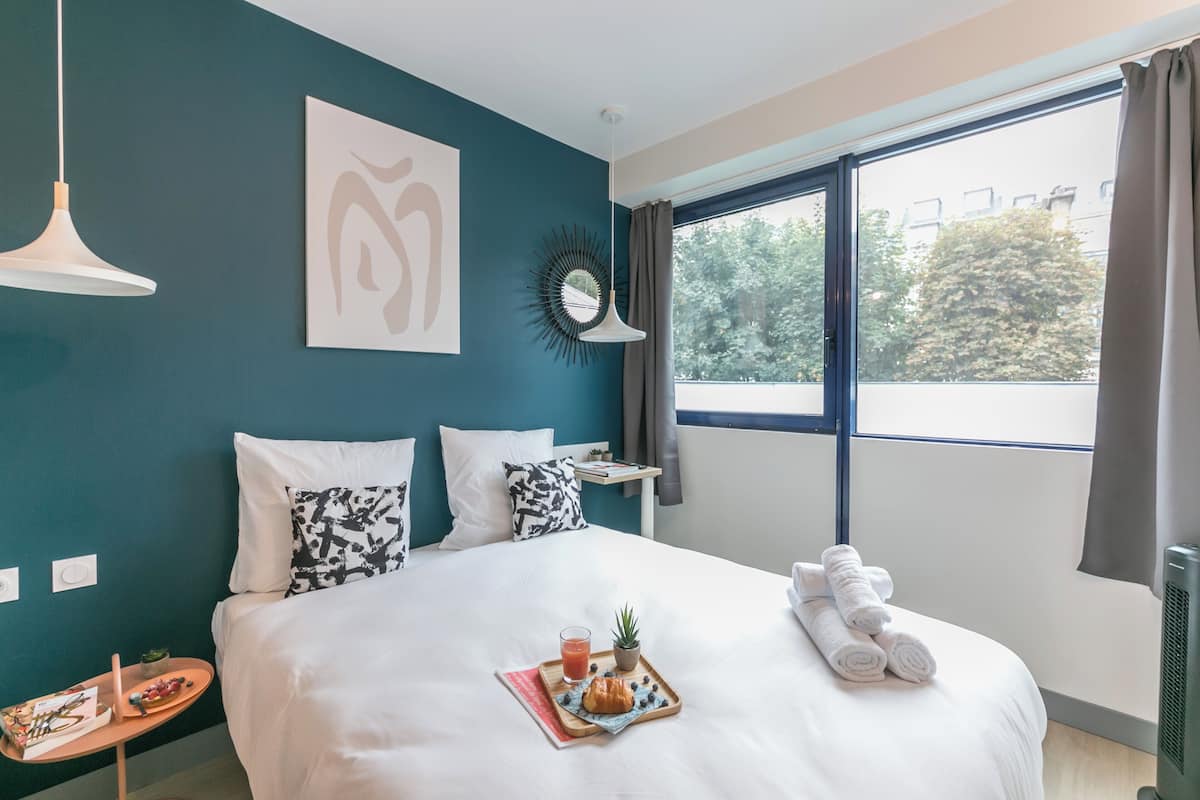 A hotel room with a bed, a tray with breakfast items, a window with curtains, and a view of trees.