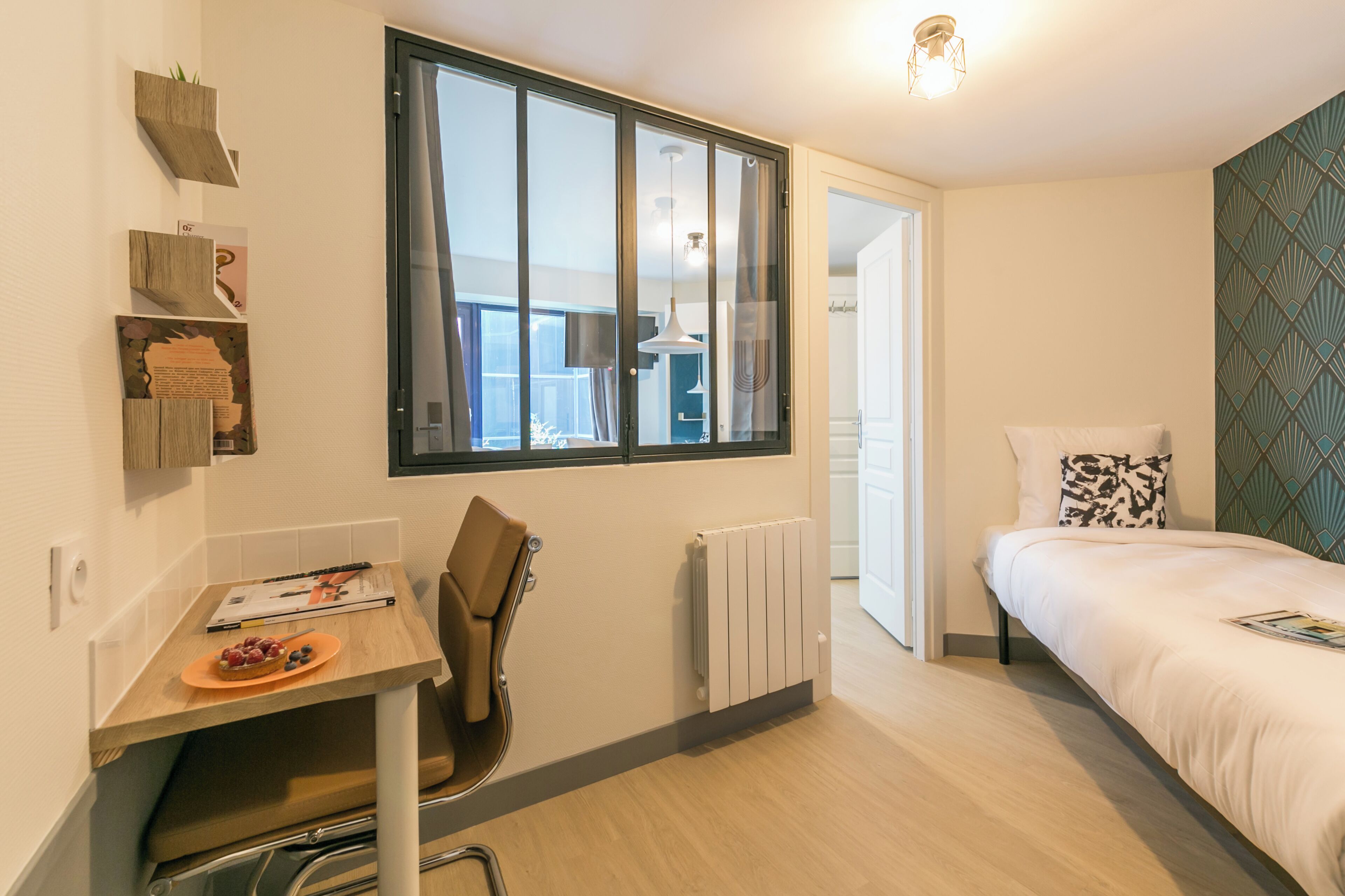 Un dormitorio pequeño con cama, escritorio con silla, radiador y ventana con vista al exterior.