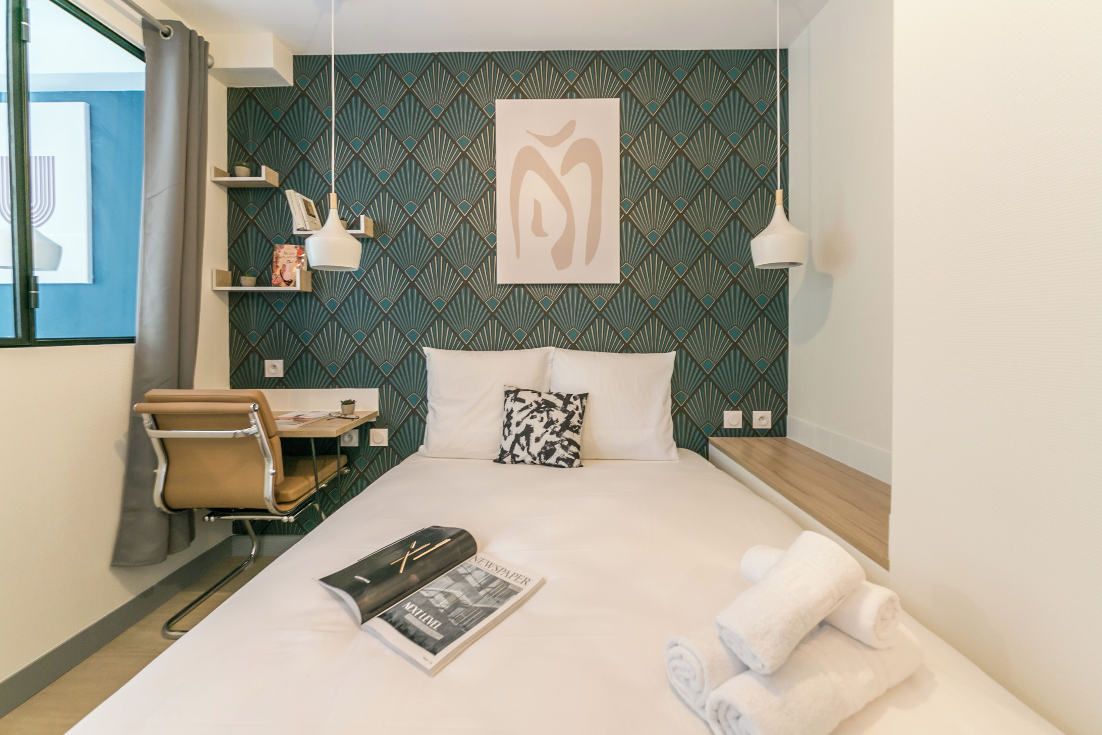 A hotel room with a bed, a desk, a chair, a window with curtains, and a wall with a geometric pattern.