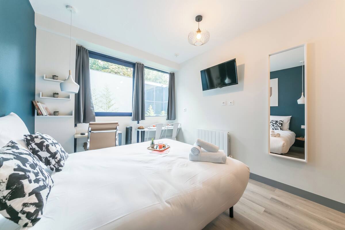 A modern hotel room with a large bed, a desk, a chair, a TV, and a window with curtains.