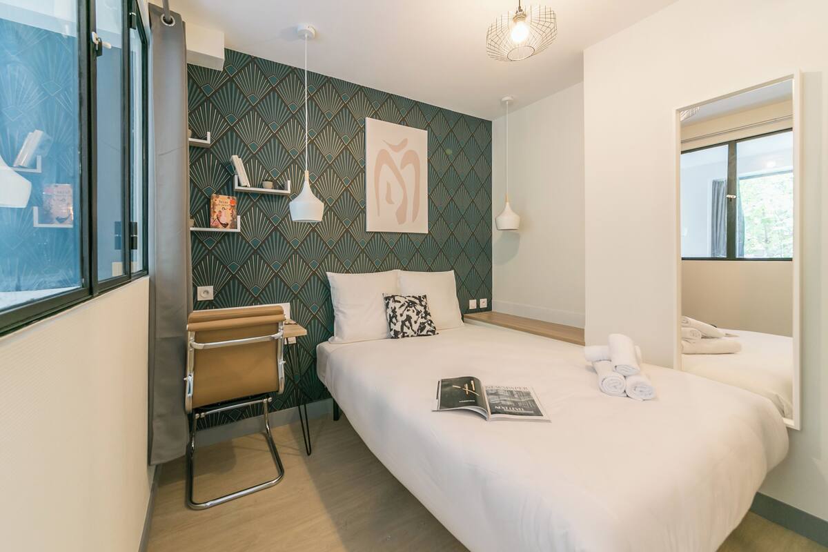 A modern hotel room with a bed, a chair, a shelf with books, a wall art piece, and a window.