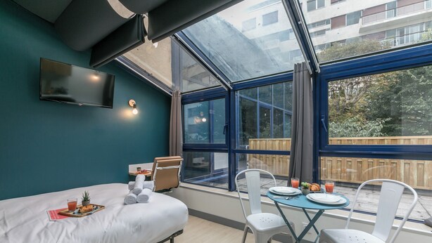 A modern room with a bed, a dining table with chairs, a TV mounted on the wall, and large windows offering a view of the outside. - WS Tour Eiffel - Nicolo (Paris)