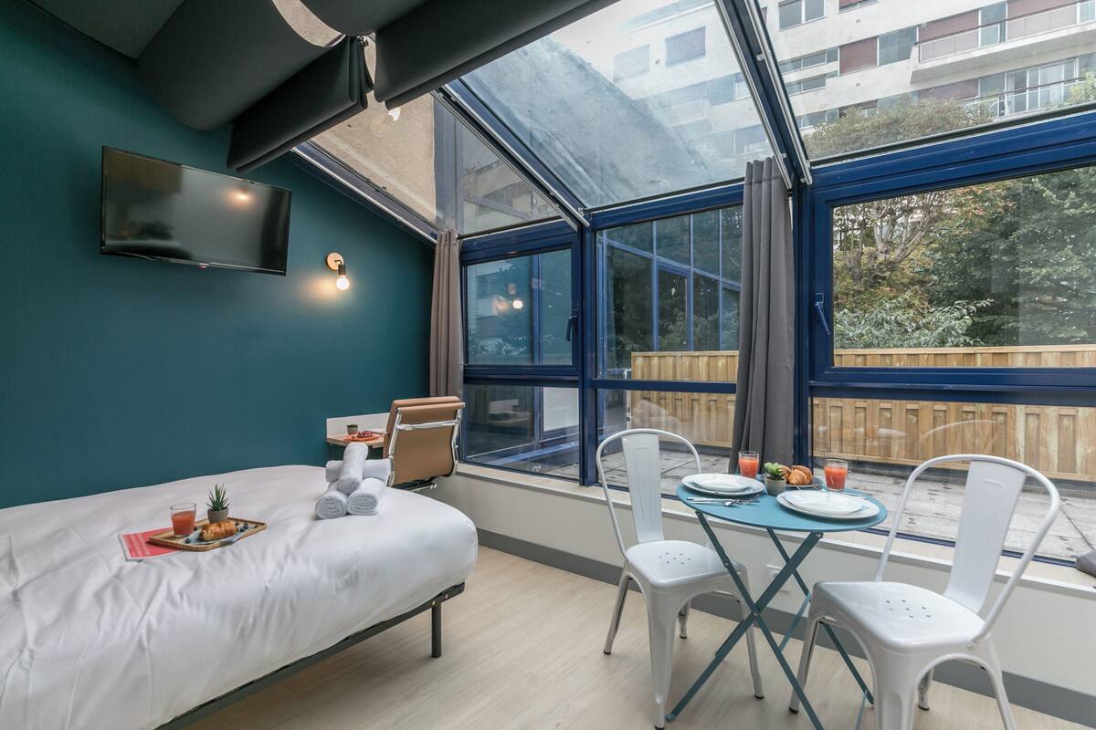 A modern room with a bed, a dining table with chairs, a TV mounted on the wall, and large windows offering a view of the outside.