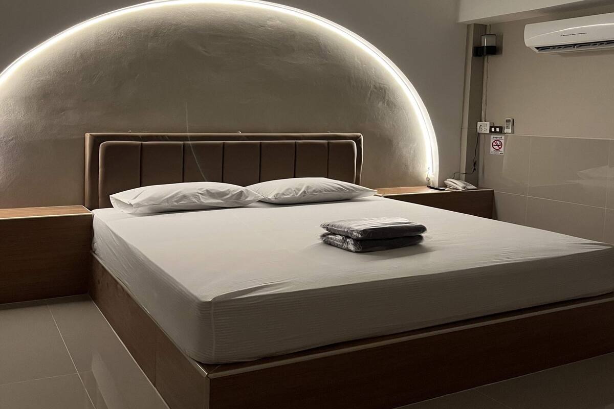 A modern hotel room with a large bed, a headboard with a curved design, and a wall-mounted air conditioning unit.