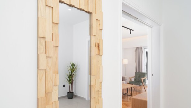 A modern room with a wooden wall panel, a potted plant, and a view into another room with a sofa and a chair.