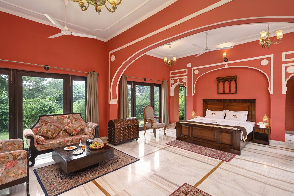 A spacious bedroom with a large bed, a sofa, a chair, and a coffee table. There are large windows and a view of greenery outside.