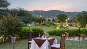 An outdoor dining area with a table set for two, surrounded by lush greenery and a view of mountains.