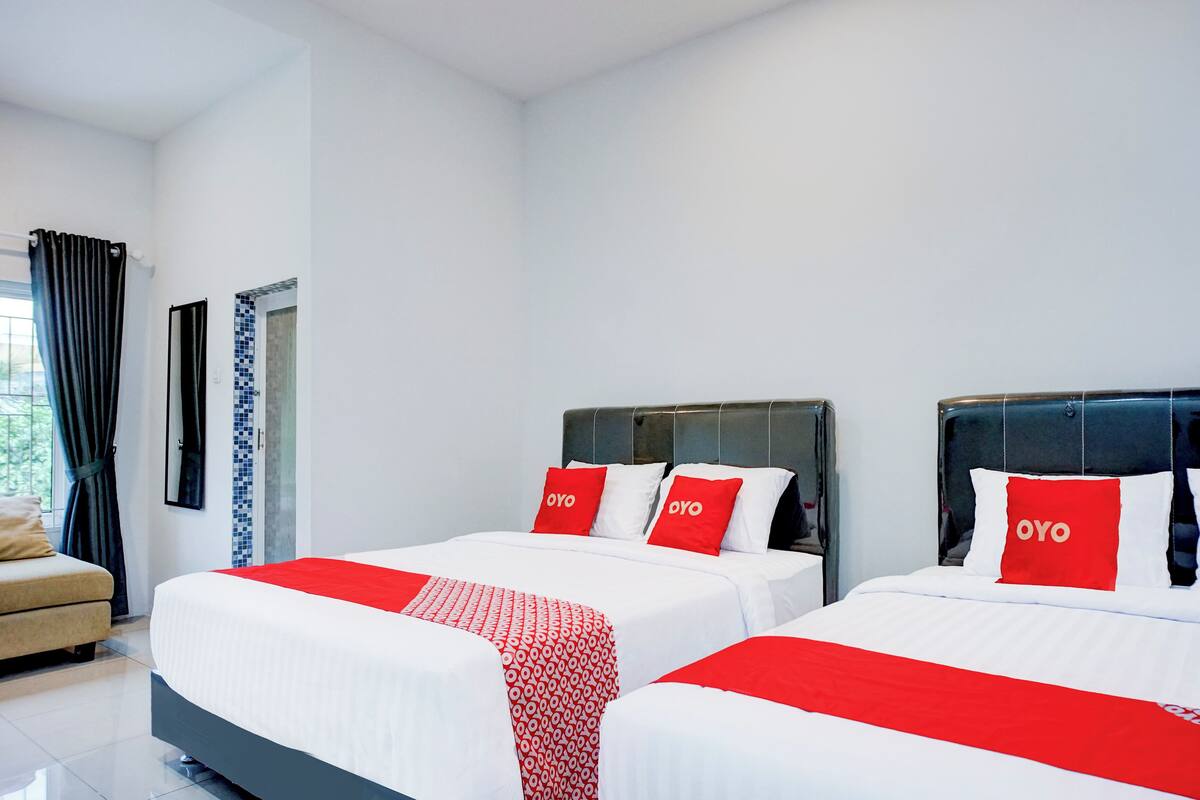 Hotellihuone, jossa on kaksi sänkyä, ja jokaisessa on punavalkoinen peitto ja tyynyjä, sekä sohva, ikkuna verhoilla ja laatoitettu seinä.