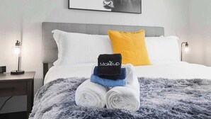 Ein ordentlich gemachtes Bett mit einer grauen, flauschigen Decke, einem weißen Kissen und einem gelben Kissen. Auf dem Bett liegen drei Handtücher gestapelt: eines schwarz mit dem Aufzug 'Mokkeup', eines blau und eines weiß. An der Wand über dem Bett hängt ein gerahmtes Bild von einem Stier.