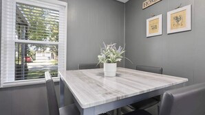 Ein Essbereich mit einem grauen Holztisch, schwarzen Stühlen, einer weißen Vase mit Blumen und einem Fenster mit Jalousien.