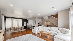A modern living room with a large sectional sofa, a wooden coffee table, and a dining area with a bench. - Beautiful Home_Large Space_3 BR_HotTub_Deck (Houston)