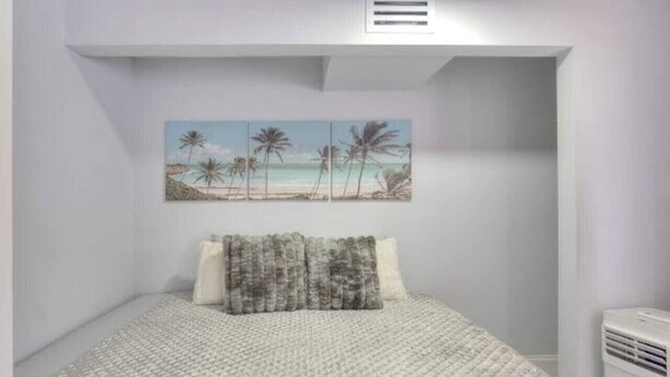 A bedroom with a bed, a painting of a beach scene, and a white air conditioning unit.