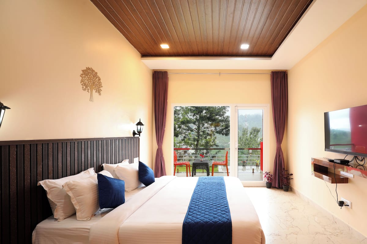 Habitación de hotel con cama, televisión, balcón con sillas rojas y vista a árboles.