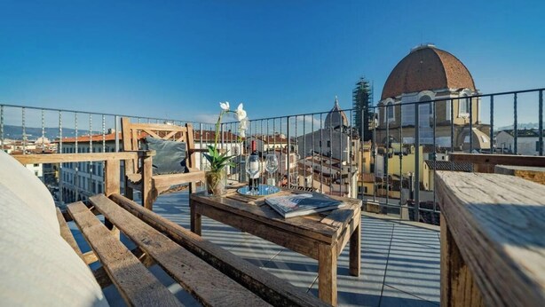 A rooftop terrace with wooden furniture, a table with a vase and glasses, and a view of a dome and buildings in the background. - Spacious 3-Bedroom Apartment with Stunning Rooftop Terraces in San Lorenzo
 (Firenze)