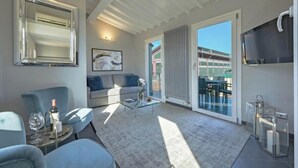 A modern living room with a glass coffee table, a sofa, a chair, a TV, and a large window with a view of a building. - Spacious 3-Bedroom Apartment with Stunning Rooftop Terraces in San Lorenzo
 (Firenze)
