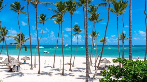 Una playa con altas palmeras, agua azul cristalina y algunas embarcaciones.
