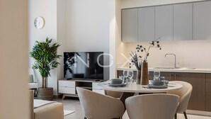 A modern living room with a flat-screen TV, a dining table with chairs, a sofa, a potted plant, and a kitchen area with cabinets and a sink. - 1BR 2510 Forte T2 (Dubai)