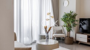 A modern living room with a sofa, a round coffee table, a chair, a television, and a large plant. - 1BR 2510 Forte T2 (Dubai)