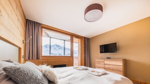 Quarto com uma cama grande, cômoda de madeira, TV de tela plana e janela com vista para as montanhas.